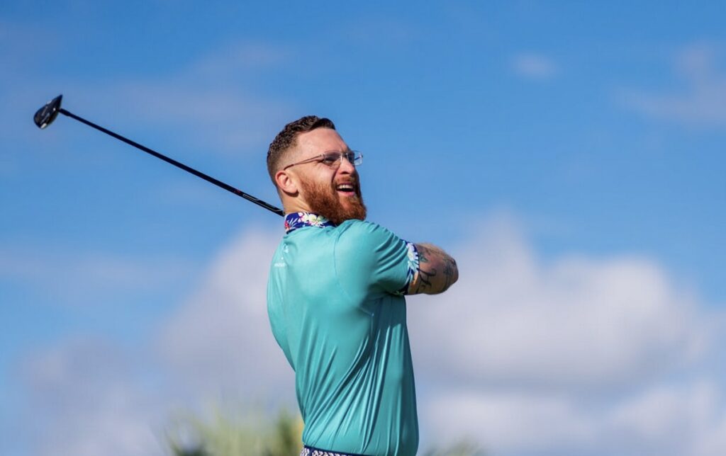 A man with a beard and glasses smiles while swinging a golf club against a clear blue sky.