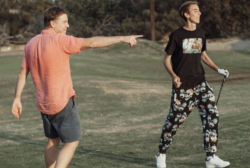 "Two people on a golf course, one in a pink shirt pointing and the other holding a golf club wearing a black t-shirt and patterned pants."