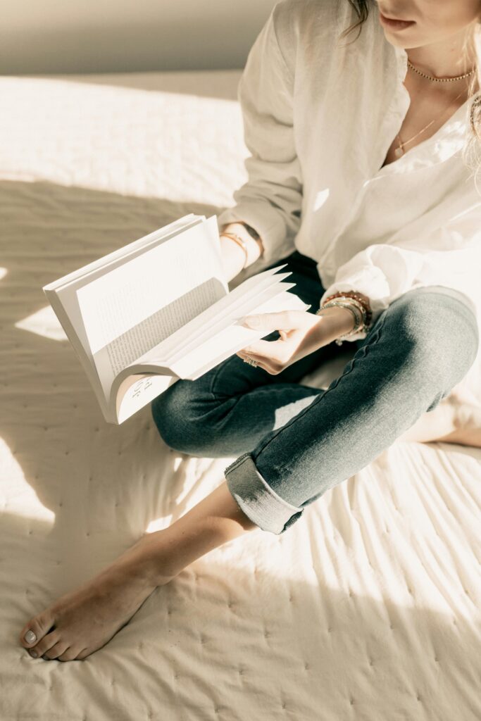 A person in a white shirt and jeans sits on a bed, reading a book in soft, natural light.