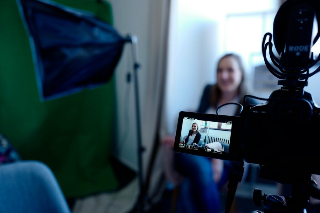 **Alt text:** A video camera recording a person sitting and smiling in a studio setup with lighting equipment.