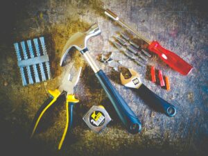 A collection of tools including a hammer, pliers, screwdriver, wrench, drill bits, wall anchors, and a tape measure, spread out on a wooden surface.