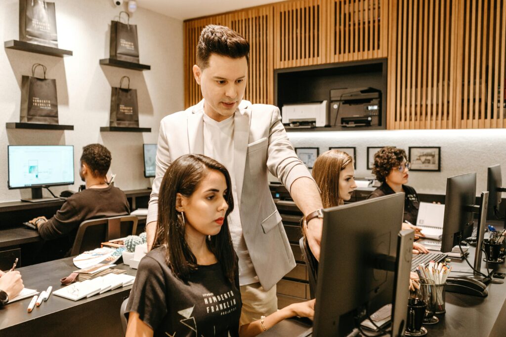 A group of people working at computer desks in an office, with a man standing and guiding a woman sitting at one of the desks.