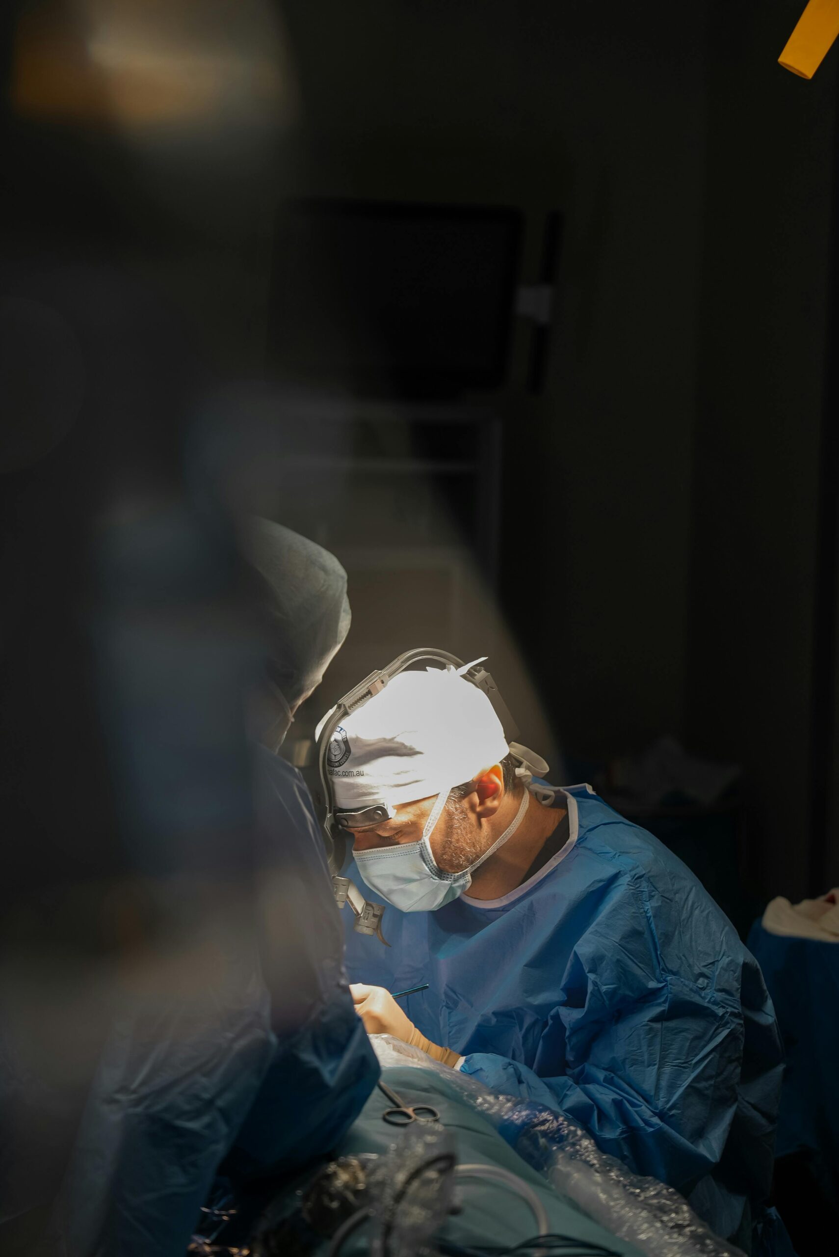 **Alt text:** A surgeon wearing surgical scrubs, a mask, and headlight performs a procedure under focused lighting in an operating room.