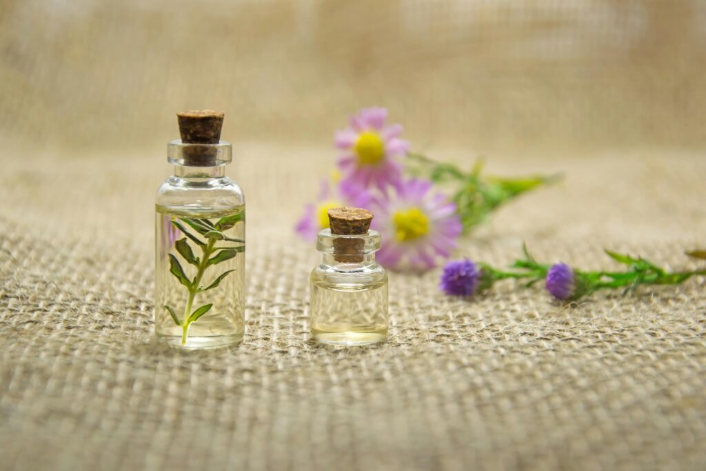 Two small glass bottles with cork stoppers, one containing a green sprig, are placed on a burlap surface, with purple and yellow flowers in the background.