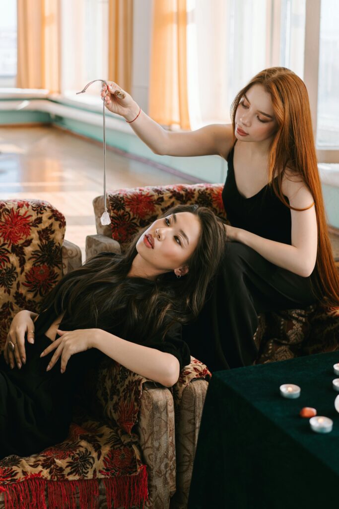 Two women are sitting on a sofa; one is lying back with closed eyes while the other holds a crystal on a string above her.