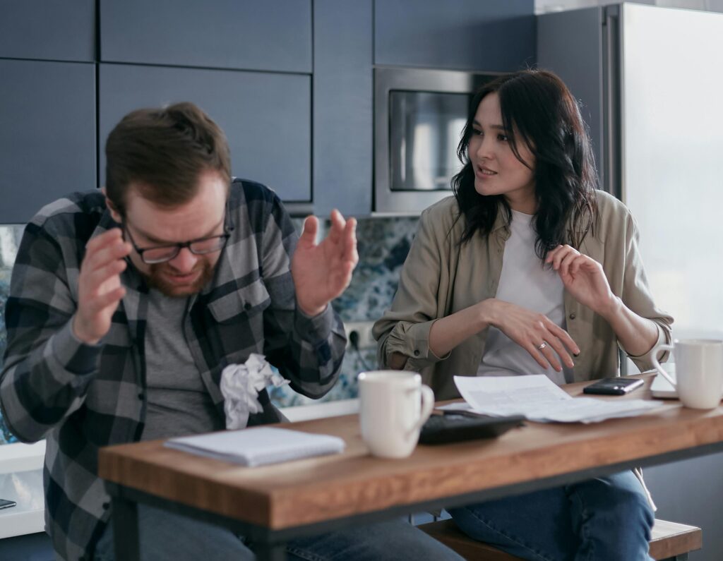 **Alt Text:** A man with glasses looks frustrated while a woman talks to him at a kitchen table with papers and mugs.
