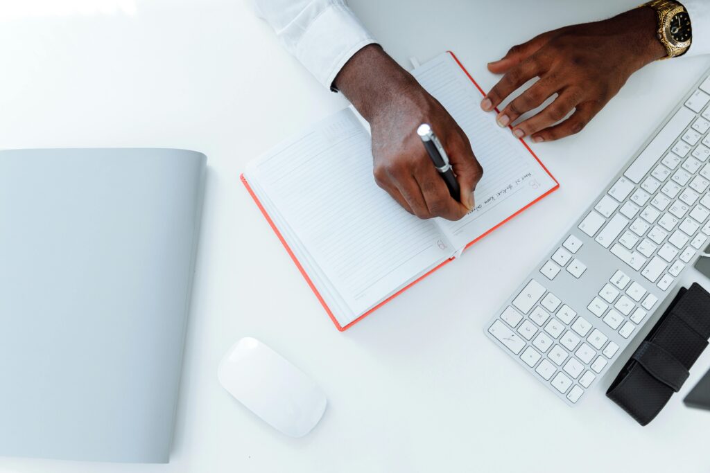 A person writing in a notebook with a red cover next to a keyboard and mouse on a white desk.