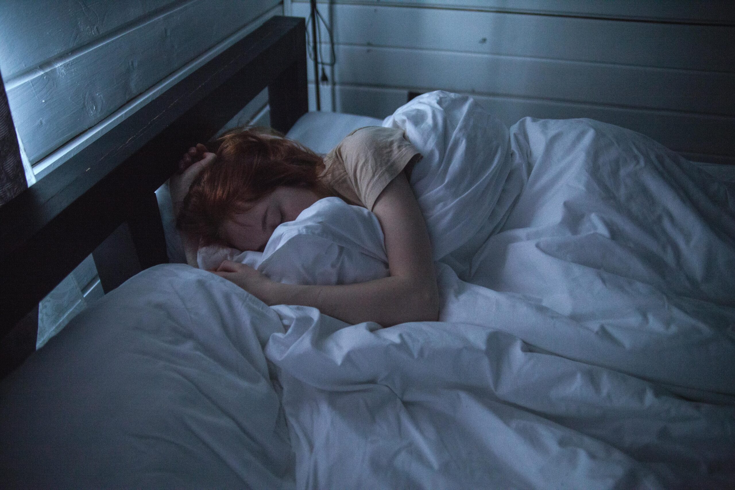 A person with red hair is sleeping in bed under a white blanket next to a wooden wall.
