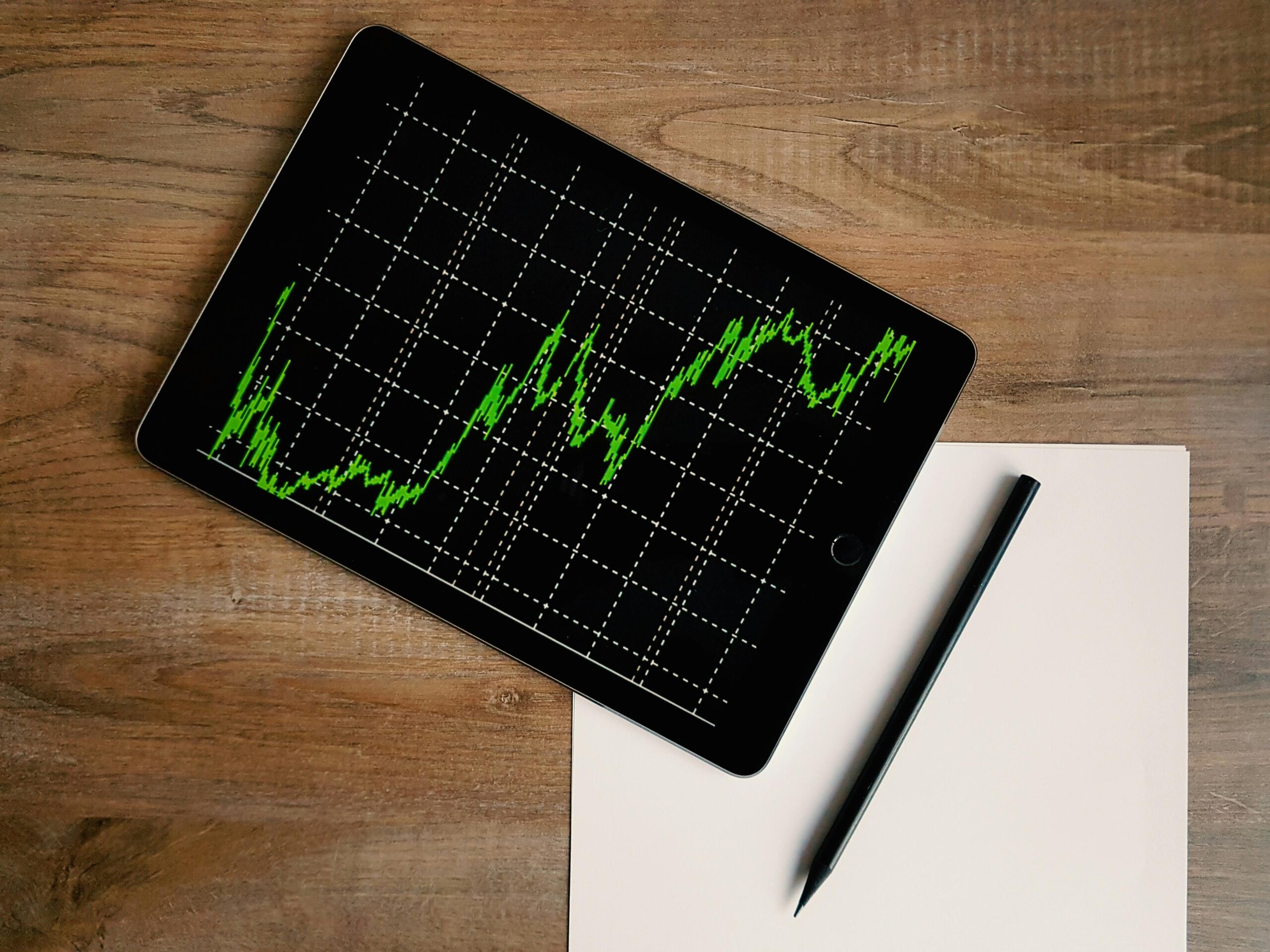 **Alt text:** A tablet displaying a fluctuating green line graph on a black background sits on a wooden table next to a blank sheet of paper and a black pen.