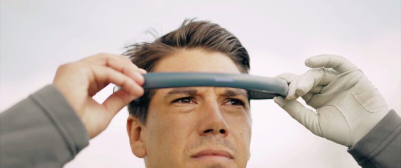 **Alt Text:** A person with short hair intently holds a gray item above their forehead, wearing gloves on their right hand.