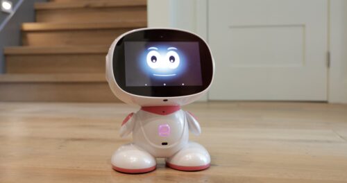 A small, white robotic device with a screen displaying a smiling face, placed on a wooden floor in front of stairs.