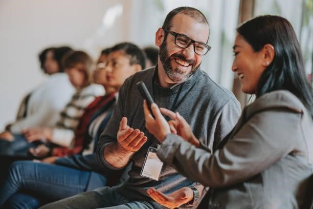 **Alt text:** A man and a woman sitting among a group, engaging in a lively conversation while looking at a phone.