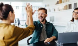 **Alt text:** A group of three people in an office setting, with one man enthusiastically high-fiving a woman across the table.