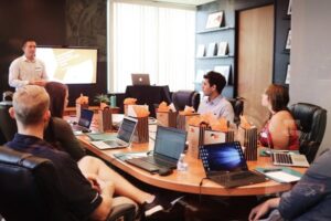<!-- Image alt text: A group of people are sitting around a conference table with laptops, watching a man present in front of a screen displaying slides. -->