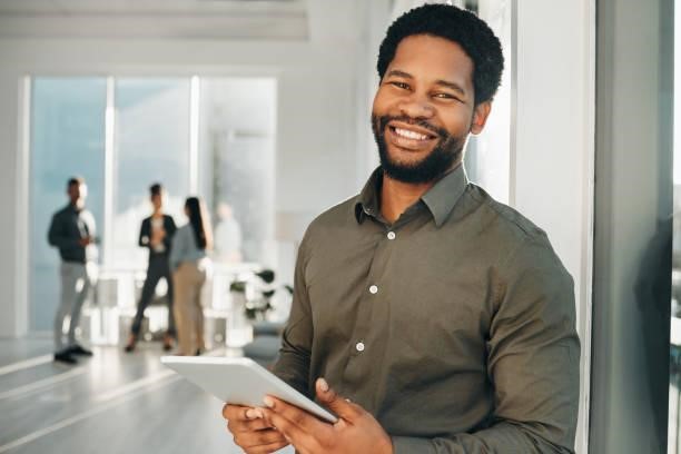 A smiling man in a dark shirt stands holding a tablet in a bright office while three people converse in the background.