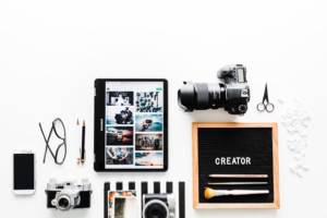 A flat lay image of various creative tools including a tablet displaying photos, a DSLR camera, glasses, pencils, paintbrushes, a smartphone, and a board with the word "CREATOR" on it.