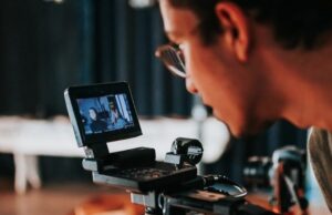 **Alt text:** A person wearing glasses looks at the screen of a professional video camera showing two people in focus.