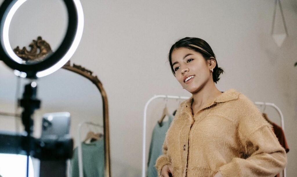 A person wearing a light brown sweater is posing with a ring light and a mirror in the background.