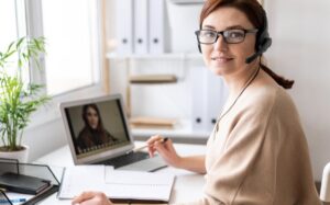 A woman wearing glasses and a headset is sitting at a desk with a laptop and taking notes during a video call.