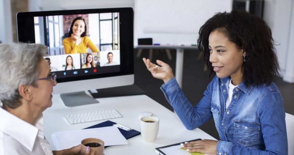 **Alt Text:** Two women are having a discussion at a desk with a computer displaying a video call with multiple participants.