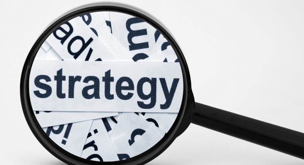 **Alt text:** A magnifying glass is placed over the word "strategy" printed on paper strips, emphasizing its focus.