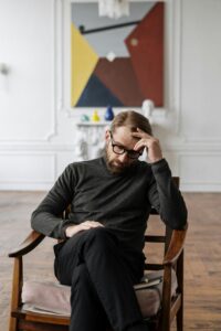 A man with glasses sits in a chair, appearing thoughtful, with his hand on his forehead, and a large abstract painting in the background.