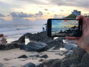 **Alt Text:** A person holds a smartphone recording a beach scene with rocks and a sunset, mirroring the view visible in the background.