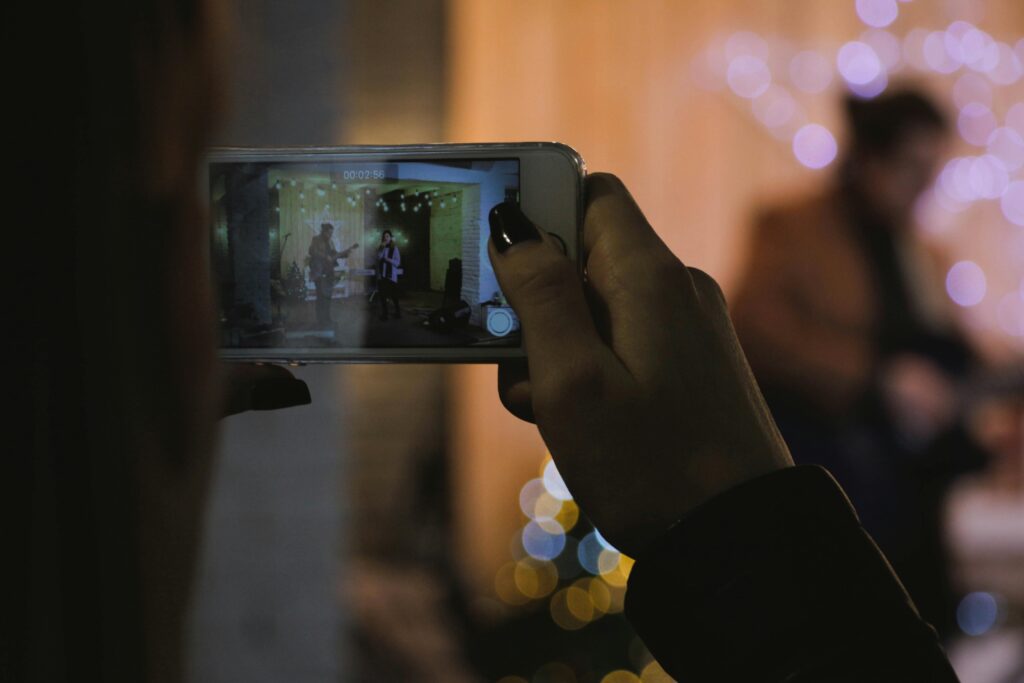 **Alt Text:** A person is recording a live performance on their phone, capturing two musicians on stage against a backdrop of string lights.