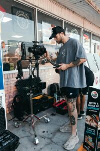 **Alt text:** A person operates a professional camera mounted on a stabilizer outside a store, with various equipment cases and a lottery advertisement nearby.