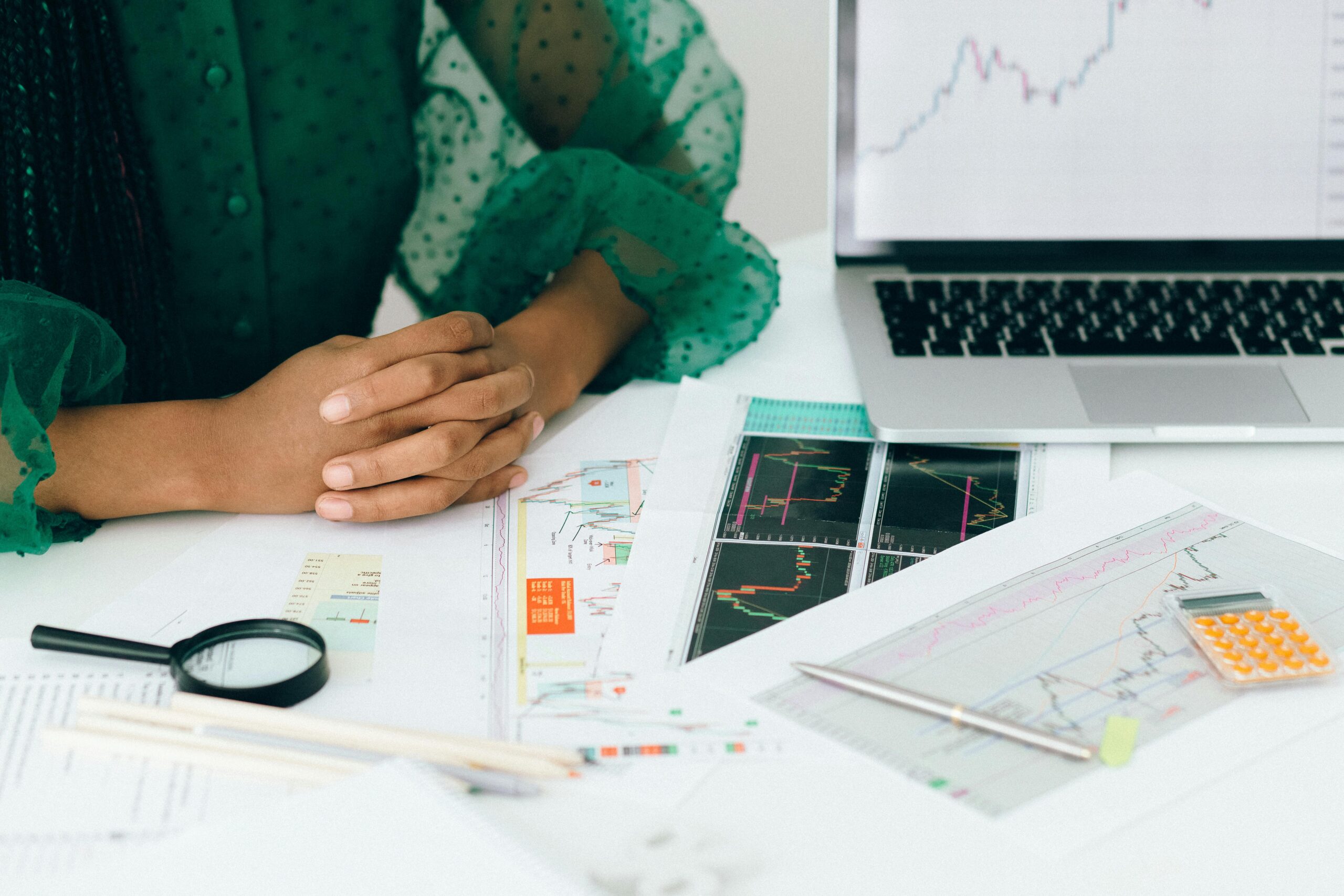 A person with clasped hands sits at a desk with financial charts, a laptop, magnifying glass, and a calculator.