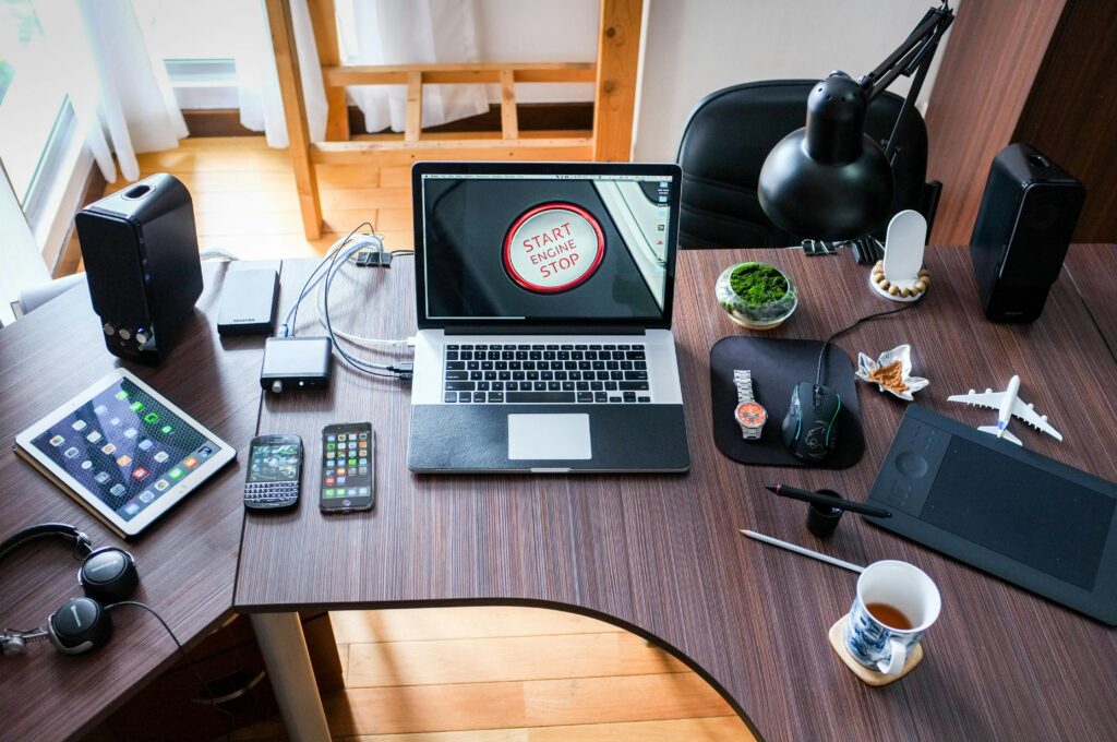 A modern workspace features a laptop with a "Start Engine Stop" screen, surrounded by smartphones, a tablet, headphones, computer accessories, and a cup of tea.
