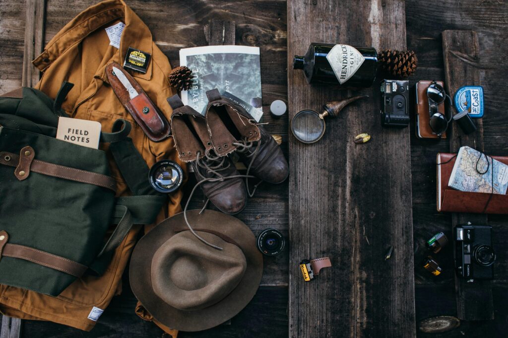 A vintage travel-themed flat lay including a jacket, leather boots, a hat, a field notes book, cameras, film, a pipe, a magnifying glass, and a bottle of gin on a wooden surface.