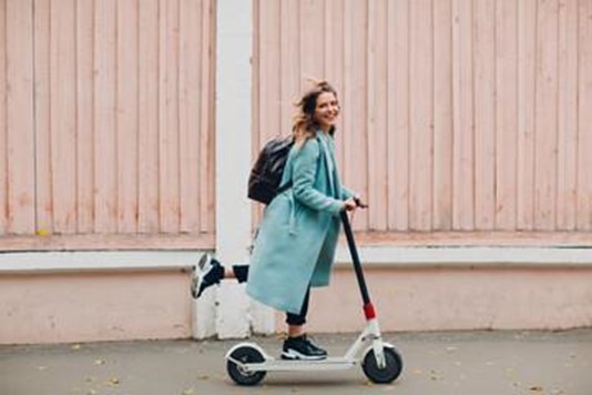 A person in a light blue coat and backpack rides a white electric scooter alongside a peach-colored wall.