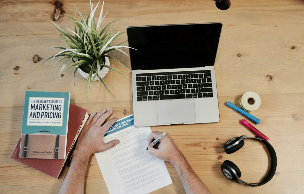 A person writes on a paper next to a laptop, a plant, a marketing book, markers, tape, and headphones on a wooden desk.