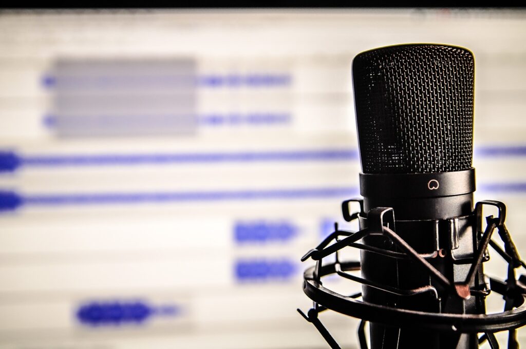 **Alt text:** A black microphone with a pop filter in focus, set against a blurred background of a computer screen displaying audio waveforms.