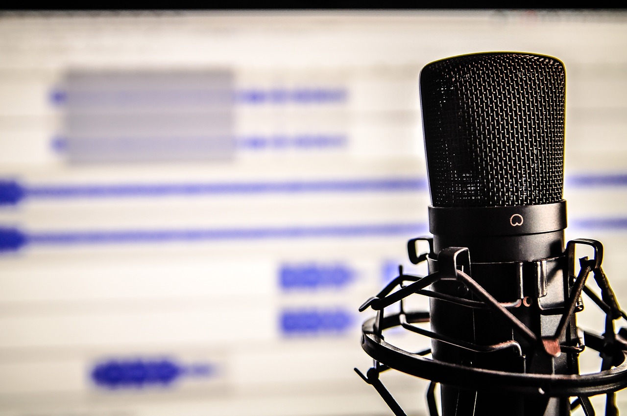 **Alt text:** A black microphone with a pop filter in focus, set against a blurred background of a computer screen displaying audio waveforms.