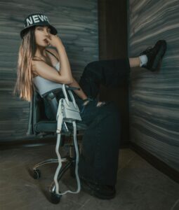 A person in a black "NEWS" hat and casual outfit sits on a chair with one leg up against a wall, holding their fingers to their lips.