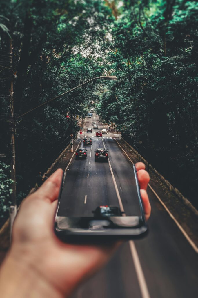 A hand holding a smartphone in front of a road lined with trees, with the phone screen seamlessly blending into the real scene of cars driving on the road.