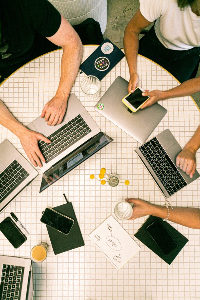 A top-down view of a group of people working at a table with laptops, smartphones, notebooks, and drinks.