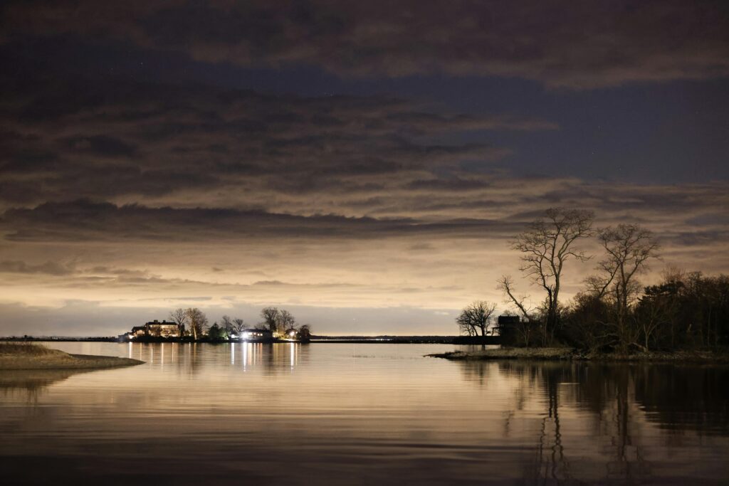 **Alt Text:** A tranquil night scene of a calm lake reflecting trees and distant lights, with a cloudy sky above.