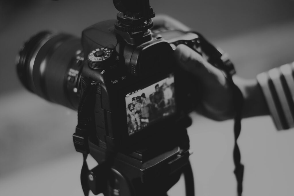 **Alt text:** A close-up black and white image of a person holding a DSLR camera, focusing on the camera screen which displays an image of people.
