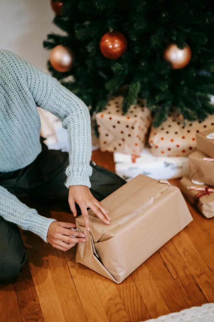 **Alt text:** A person in a knitted sweater wraps a gift in brown paper near decorated Christmas tree.