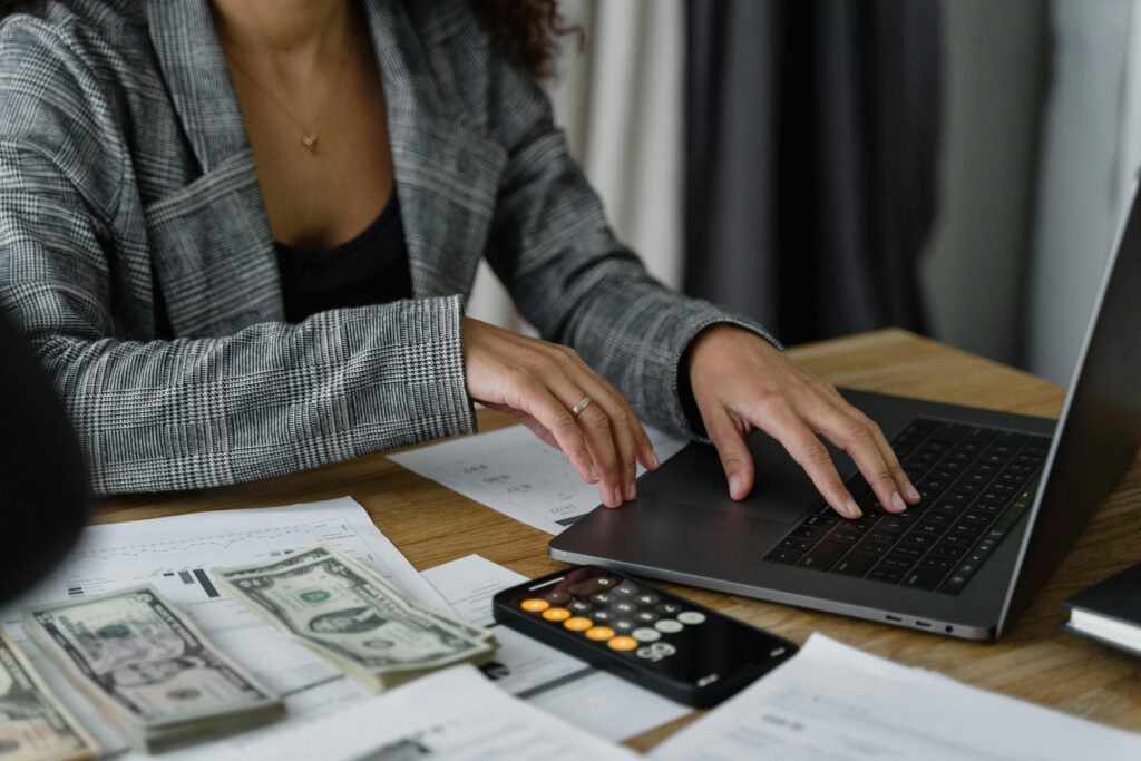 A person in a gray plaid jacket uses a laptop on a desk with scattered money, documents, and a smartphone calculator.