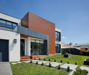 A modern house with a flat roof, large windows, and a combination of white, gray, and wooden exterior elements, set against a blue sky.