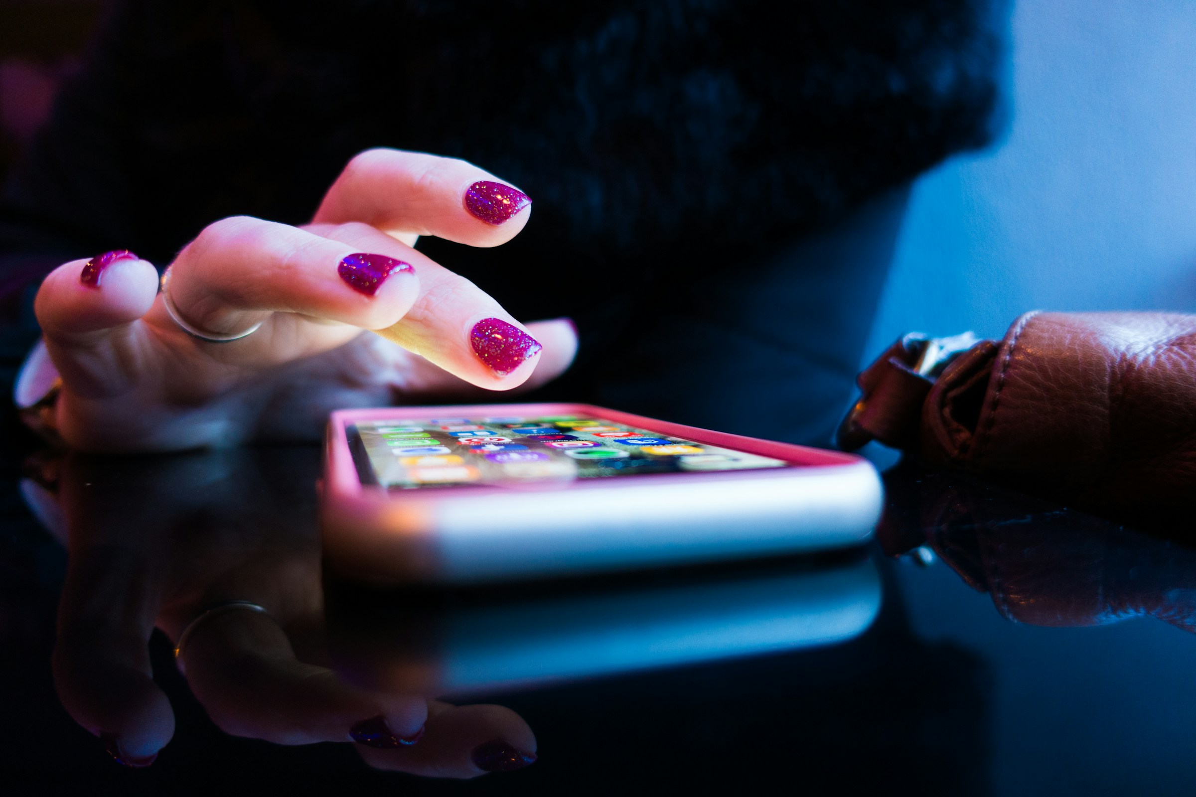 **Alt Text:** A hand with sparkly purple nail polish is tapping on a smartphone screen on a reflective surface.