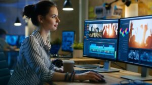 A woman in a striped shirt works on video editing at a dual-monitor setup, displaying video clips and timelines.