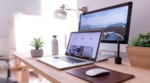 A clean and modern desk setup with a computer monitor displaying "Work Hard Anywhere," a laptop, a plant, a water bottle, and a mouse on a leather pad.