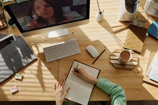 A person writes in a notebook on a wooden desk while participating in a video call on a computer screen.