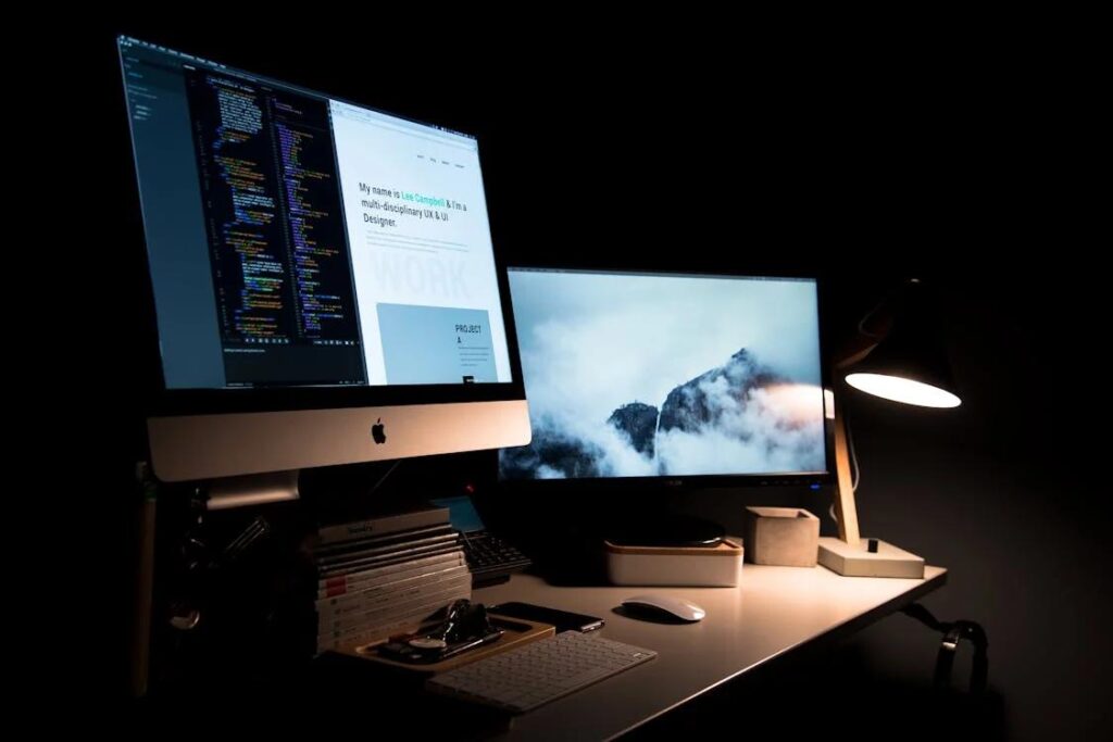 A dimly lit workspace with dual monitors displaying code and a mountain landscape, alongside a keyboard, mouse, and desk lamp.