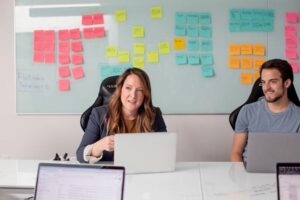 **Alt Text:** Two people sitting at a table with laptops, in front of a whiteboard covered in colorful sticky notes.